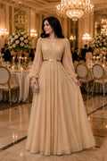 A woman stands in a grand event hall wearing a floor-length champagne beige gown with long sheer sleeves, a V-neckline, and a thin metallic belt at the waist. The background features elegant dining tables, floral arrangements, and large chandeliers.