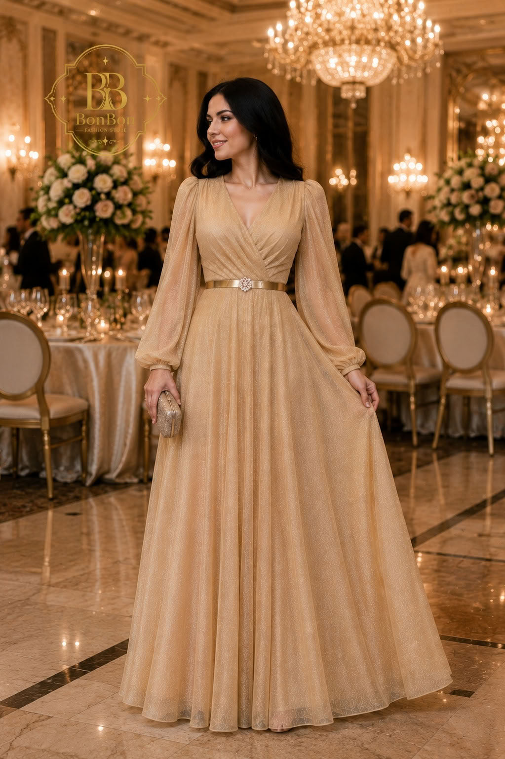 A woman stands in a grand event hall wearing a floor-length champagne beige gown with long sheer sleeves, a V-neckline, and a thin metallic belt at the waist. The background features elegant dining tables, floral arrangements, and large chandeliers.