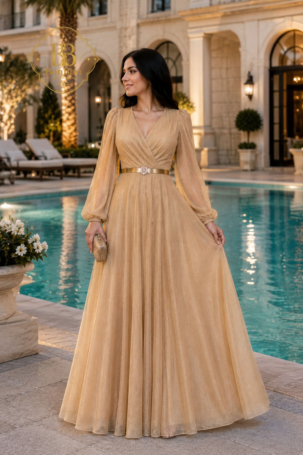 A woman wearing a floor-length champagne beige gown with long sheer sleeves and a gold belt stands by a luxury resort swimming pool at dusk.
