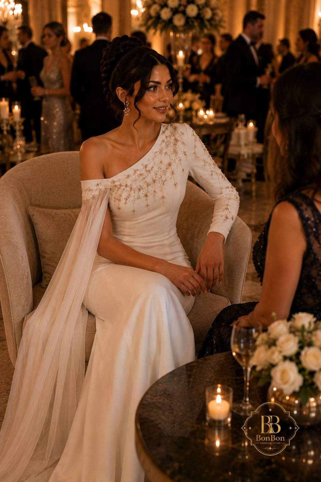 A woman seated in a formal event hall wearing an off-the-shoulder white gown with long sleeves and delicate gold embellishments, with a long train draped over a chair. Guests and tables with candles are visible in the warm, dimly lit background.