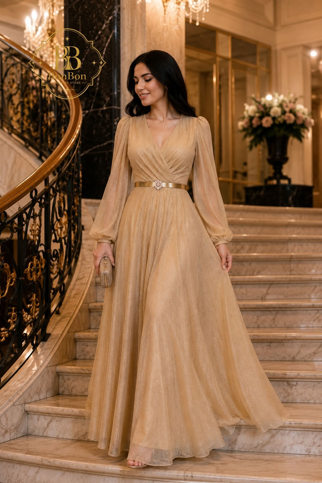 A woman wearing a floor-length champagne beige gown with long sheer sleeves and a metallic gold belt, standing on marble stairs in an elegant indoor setting.