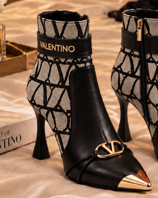 A pair of Valentino pointed-toe ankle boots with black leather, a gray geometric pattern, and gold-tone hardware, displayed on a bed with satin bedding and dark velvet fabric.
