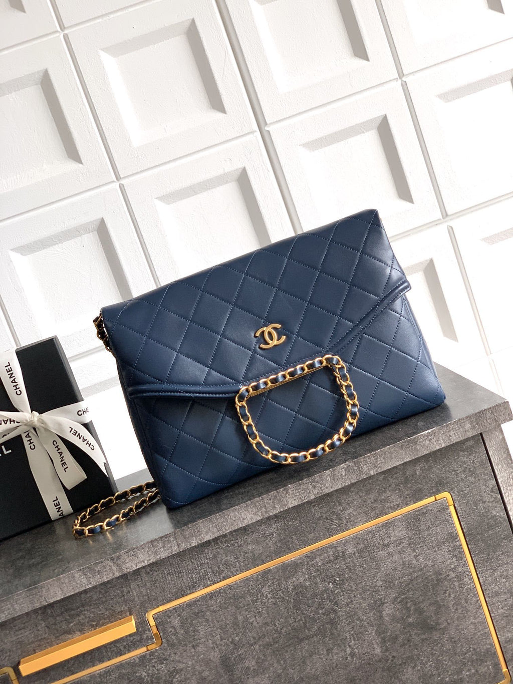 Blue quilted handbag with gold chain on a gray surface with white geometric patterned background