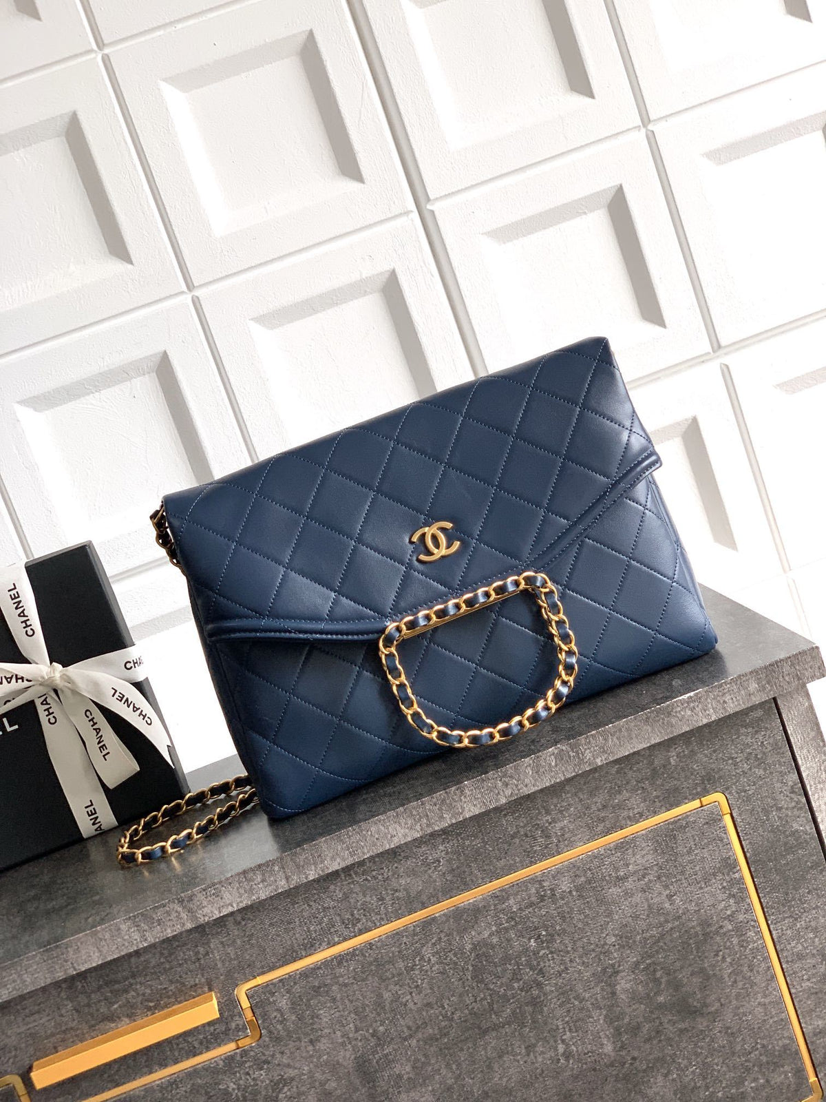 Blue quilted handbag with gold chain on a gray surface with white geometric patterned background