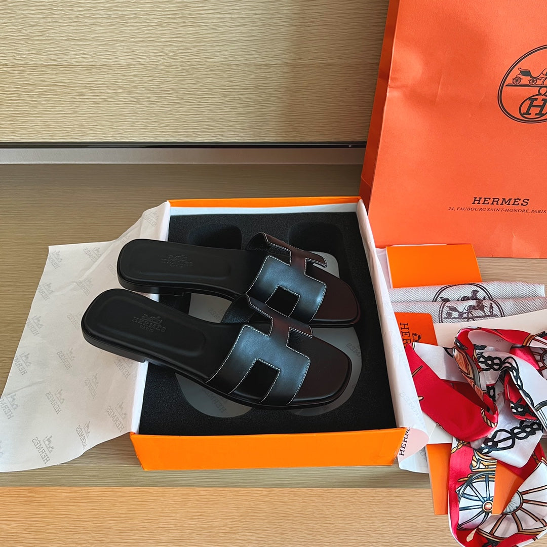 A pair of black leather Hermes slide sandals with signature H-strap details, presented in their orange and white box alongside an orange Hermes shopping bag and a patterned scarf on a wooden surface.