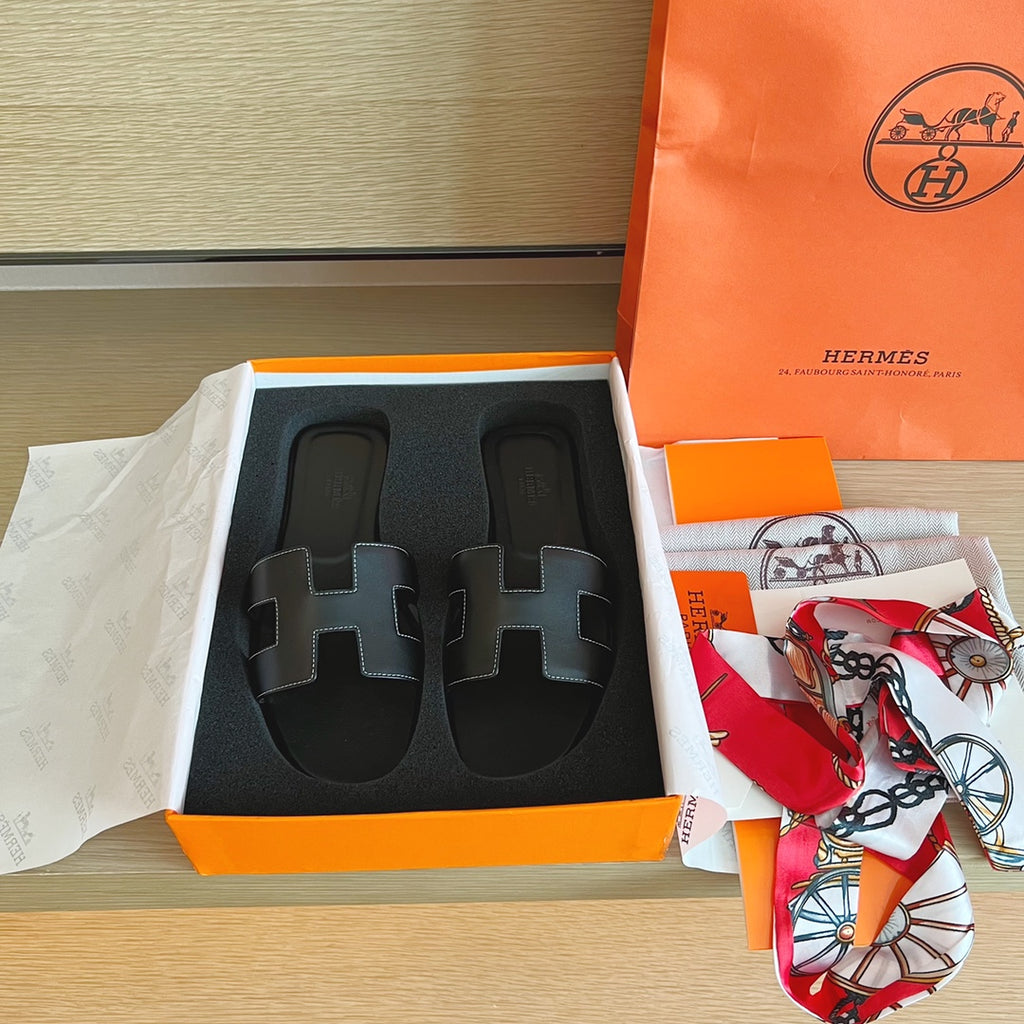 A pair of black leather Hermes H-slide sandals in their original orange and white box, with an orange Hermes shopping bag and a patterned silk scarf on a light wood table.
