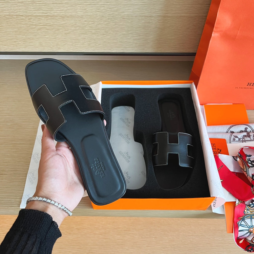 A hand holds a black leather Hermès H slide sandal next to its orange and white box on a wooden table, with an orange shopping bag and accessories visible in the background.