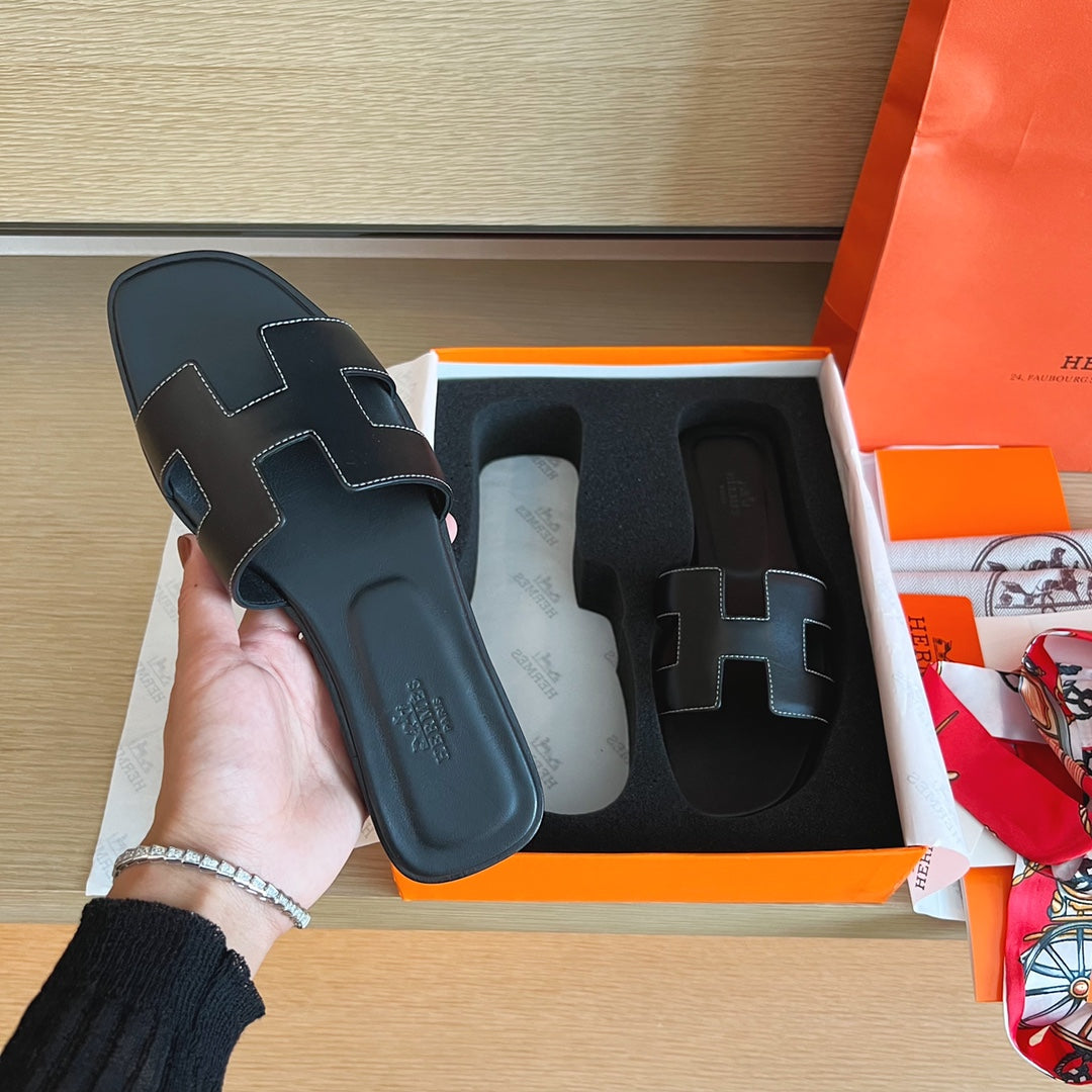 A hand holds a black leather Hermès H slide sandal next to its orange and white box on a wooden table, with an orange shopping bag and accessories visible in the background.