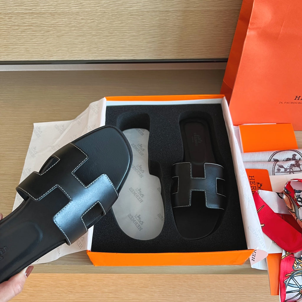 A black leather Hermès H-strap slide sandal held by a hand, positioned next to its orange and white Hermès box on a wooden surface.