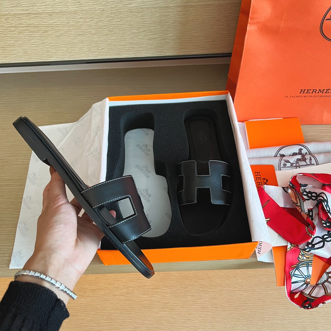 A hand holds a black leather Hermes slide sandal next to its orange and white box, with another sandal in the foam insert and an orange Hermes shopping bag on a wooden table.