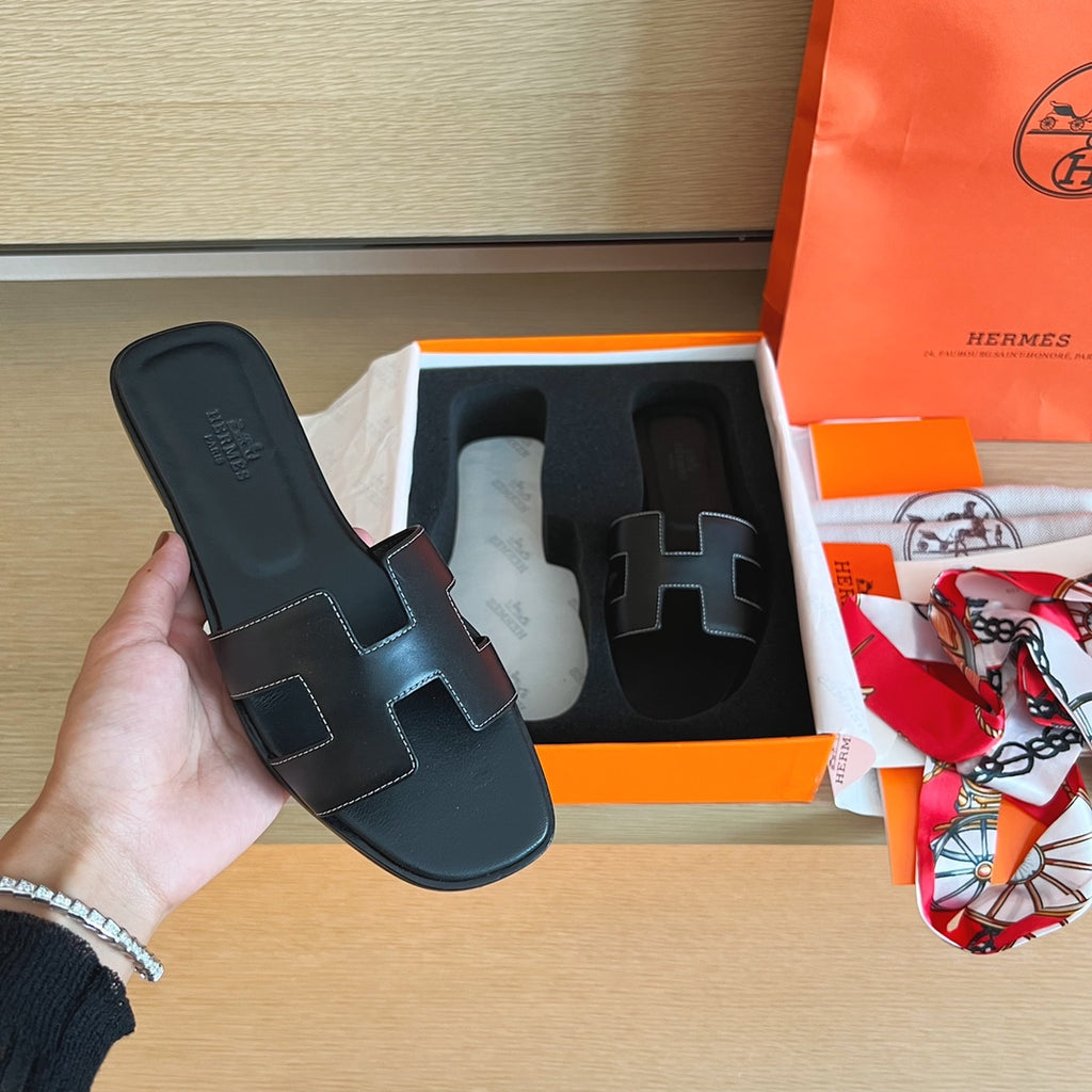 A hand holds a black leather Hermes H-slide sandal next to its orange and white box, with an orange Hermes shopping bag and a patterned scarf on a light wood surface.