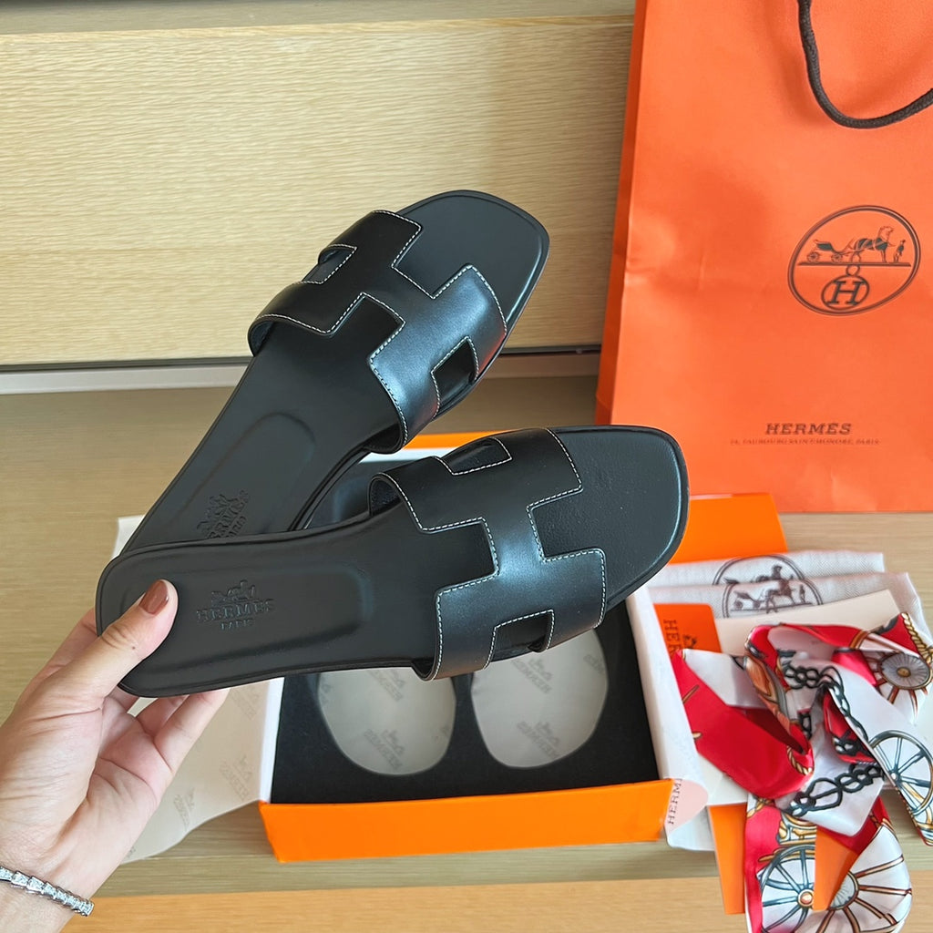 A pair of black leather Hermès slide sandals with signature cutout straps, held by a hand next to an orange Hermès shopping bag, box, and scarf on a wooden surface.