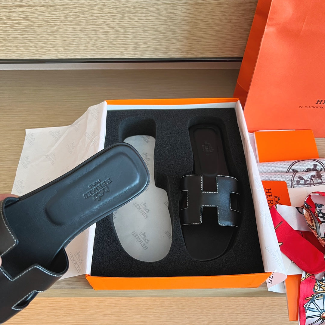 A black leather Hermes slide sandal with an H-strap is shown next to its orange and white Hermes box, with a branded shopping bag visible in the background.