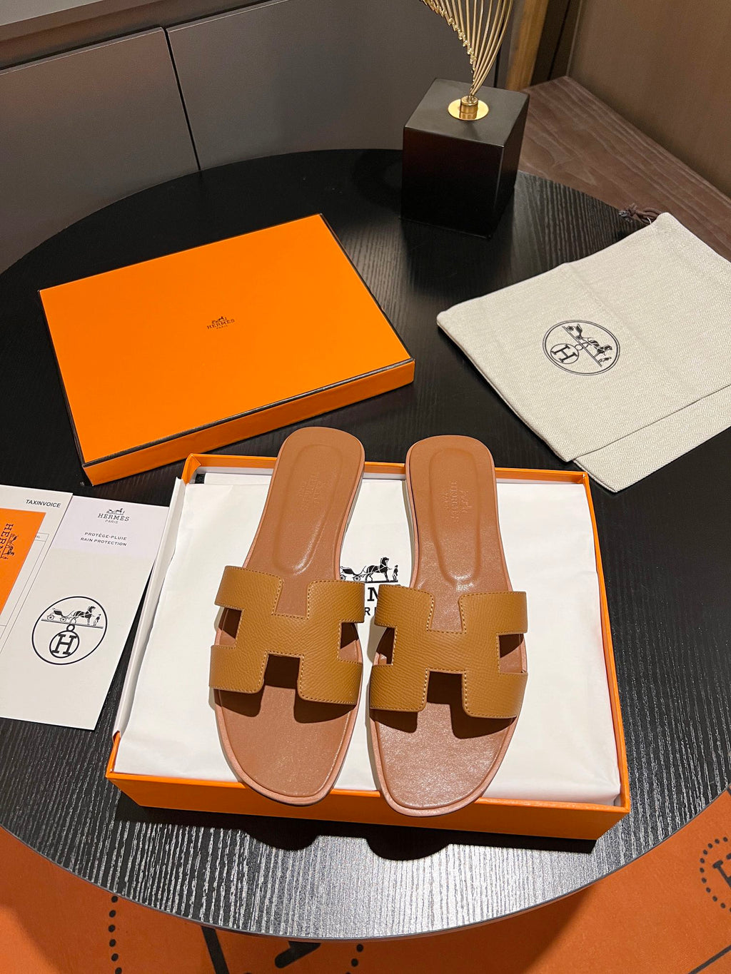 A pair of classic orange-boxed Hermès H-strap slide sandals in tan leather, displayed on a dark round table alongside the orange packaging box, dust bag, and papers.