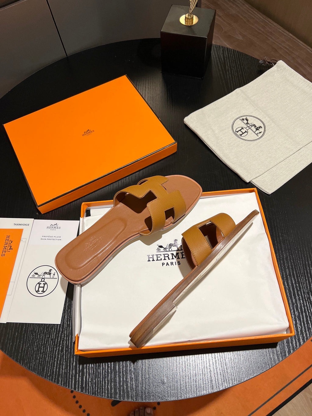 A pair of tan leather Hermès slide sandals displayed in their original orange and white packaging on a dark round table.