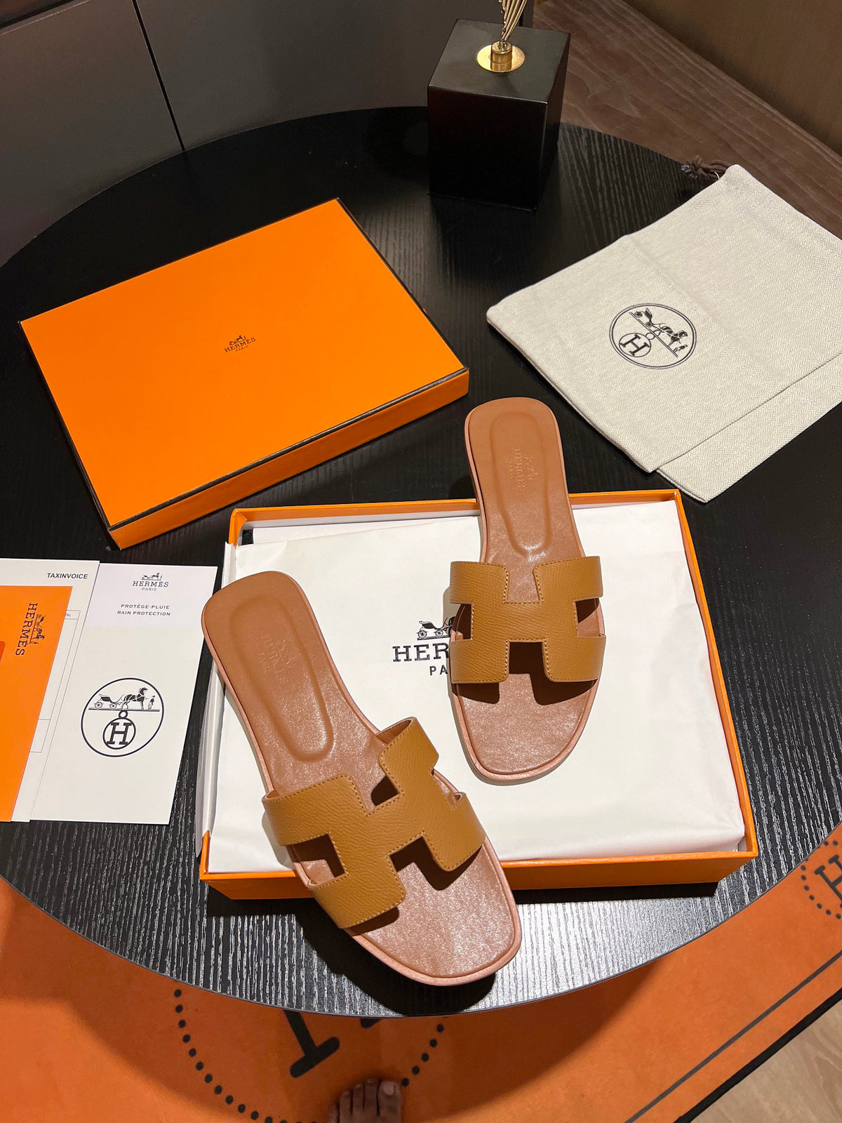 Brown sandals with gold 'H' logo on a black surface with an orange box and white towel.