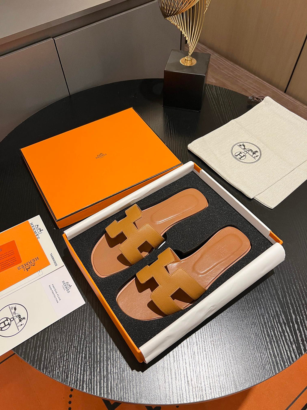 A pair of Hermès H-strap slide sandals in tan leather, presented in an open orange and white box with an orange dust bag and papers on a dark round table.