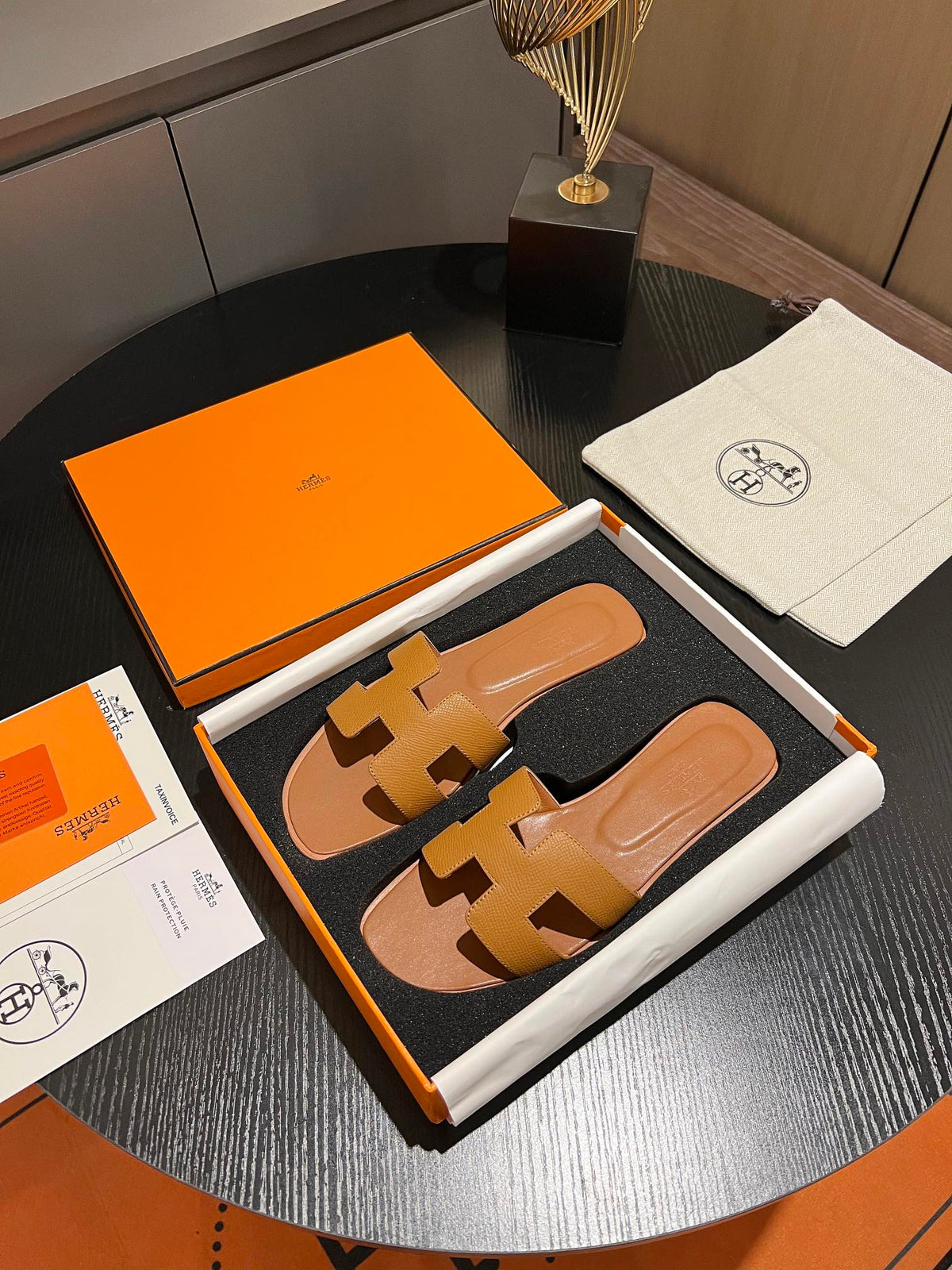 A pair of Hermès H-strap slide sandals in tan leather, presented in an open orange and white box with an orange dust bag and papers on a dark round table.