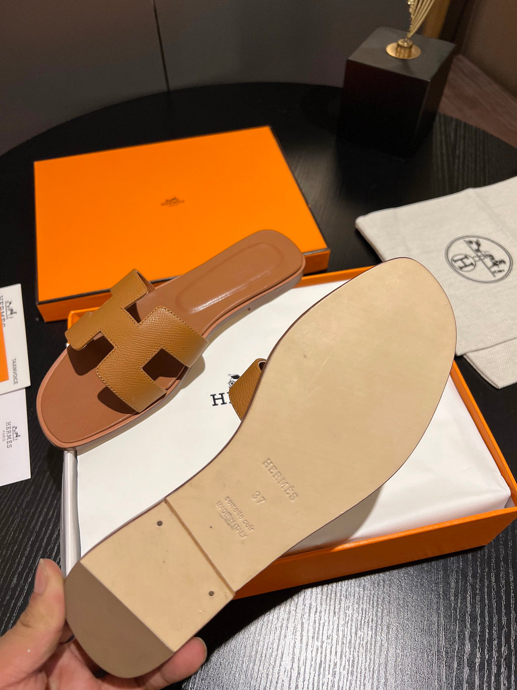 A tan leather H-strap slide sandal shown with its orange and white Hermès packaging on a dark table, with a hand holding the sandal's sole.