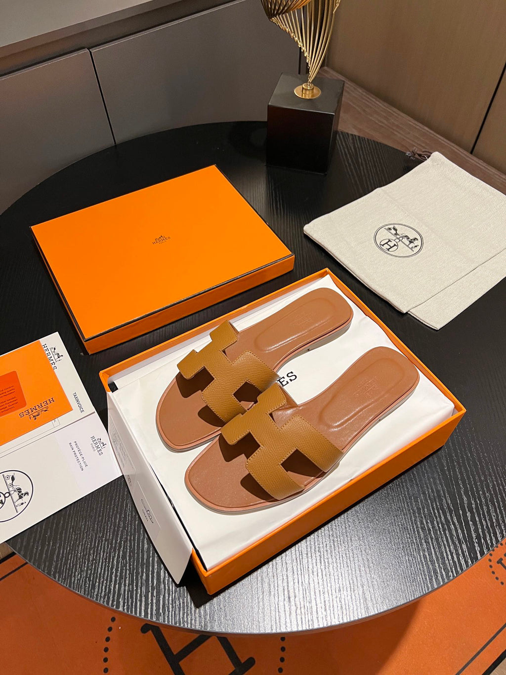 A pair of tan leather slide sandals with an H-shaped strap, presented in an orange and white box on a dark round table, alongside an orange box lid, a white dust bag with the Hermes logo, and papers.