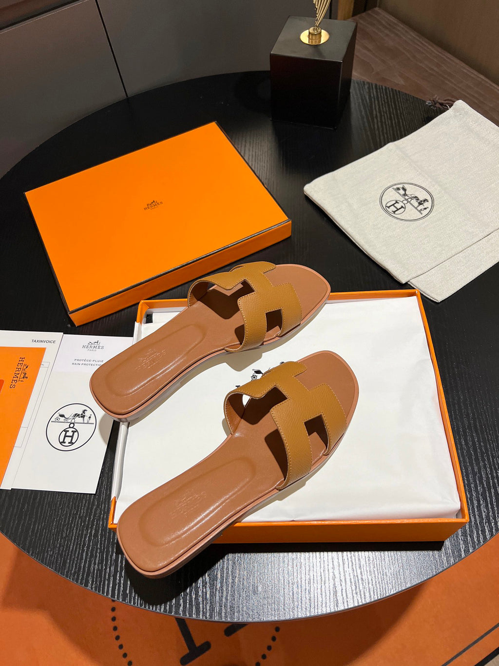 A pair of Hermès H-strap slide sandals in tan leather, presented in their original orange and white packaging on a dark round table.