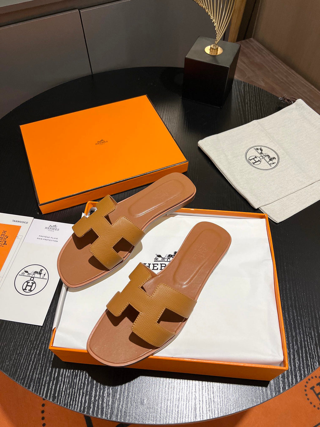 A pair of Hermès H-strap slide sandals in tan leather, presented in their signature orange box with a white dust bag and packaging cards on a dark round table.