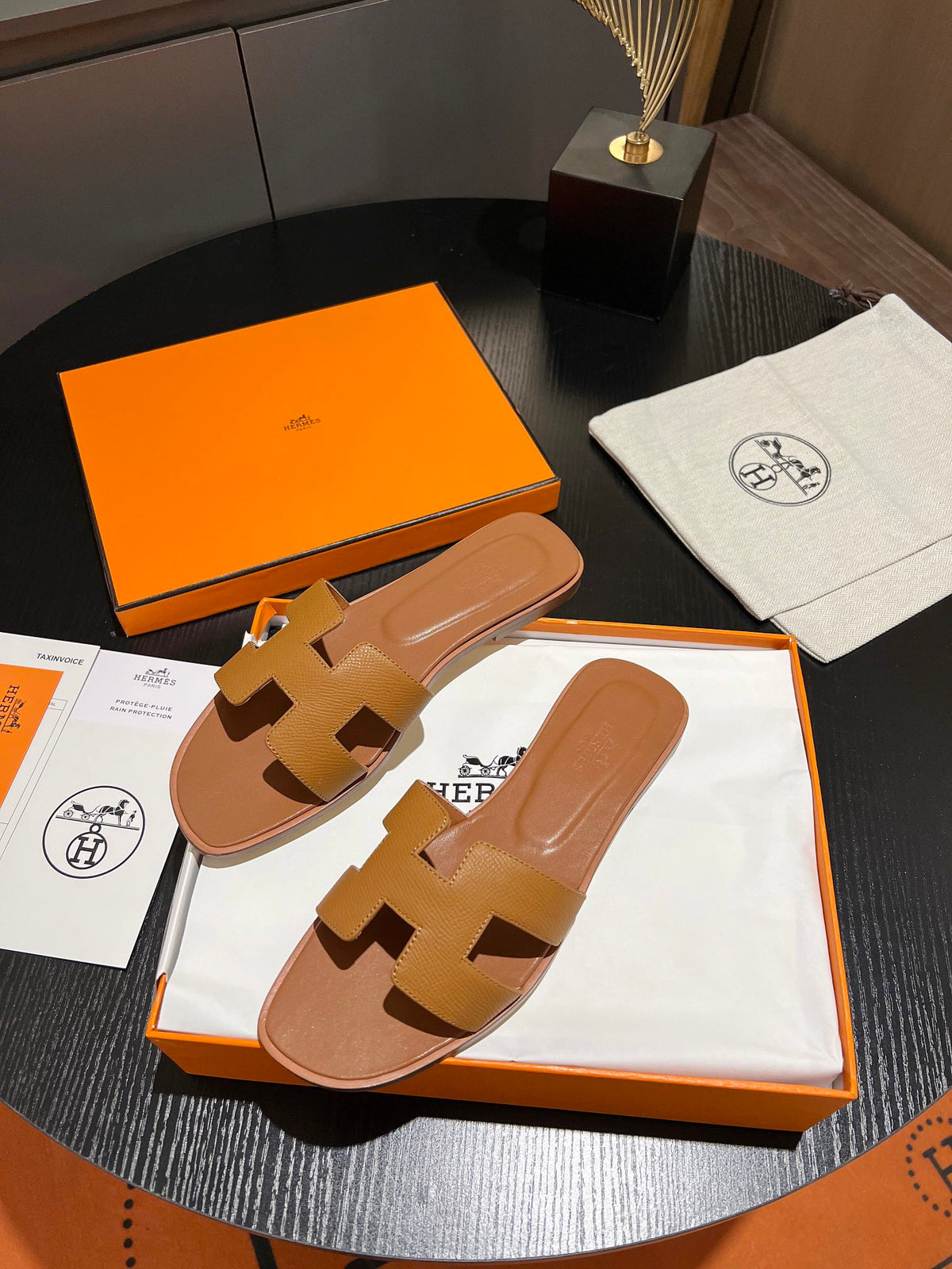 A pair of Hermès H-strap slide sandals in tan leather, presented in their signature orange box with a white dust bag and packaging cards on a dark round table.