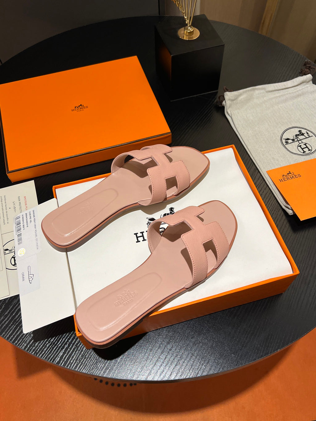 A pair of pink leather slide sandals with the signature H-strap, displayed on their orange and white Hermes packaging on a dark round table.