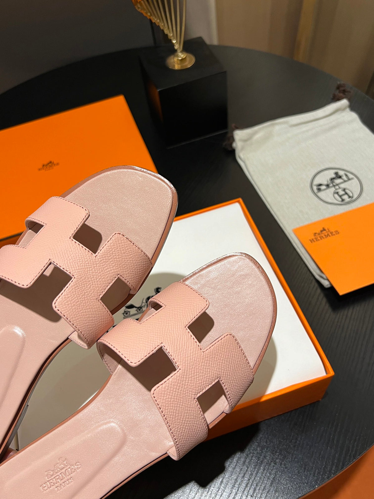 A pair of pink leather slide sandals with the signature H-shaped strap, displayed on a dark table with an orange and white Hermès box, a branded dust bag, and an orange card.