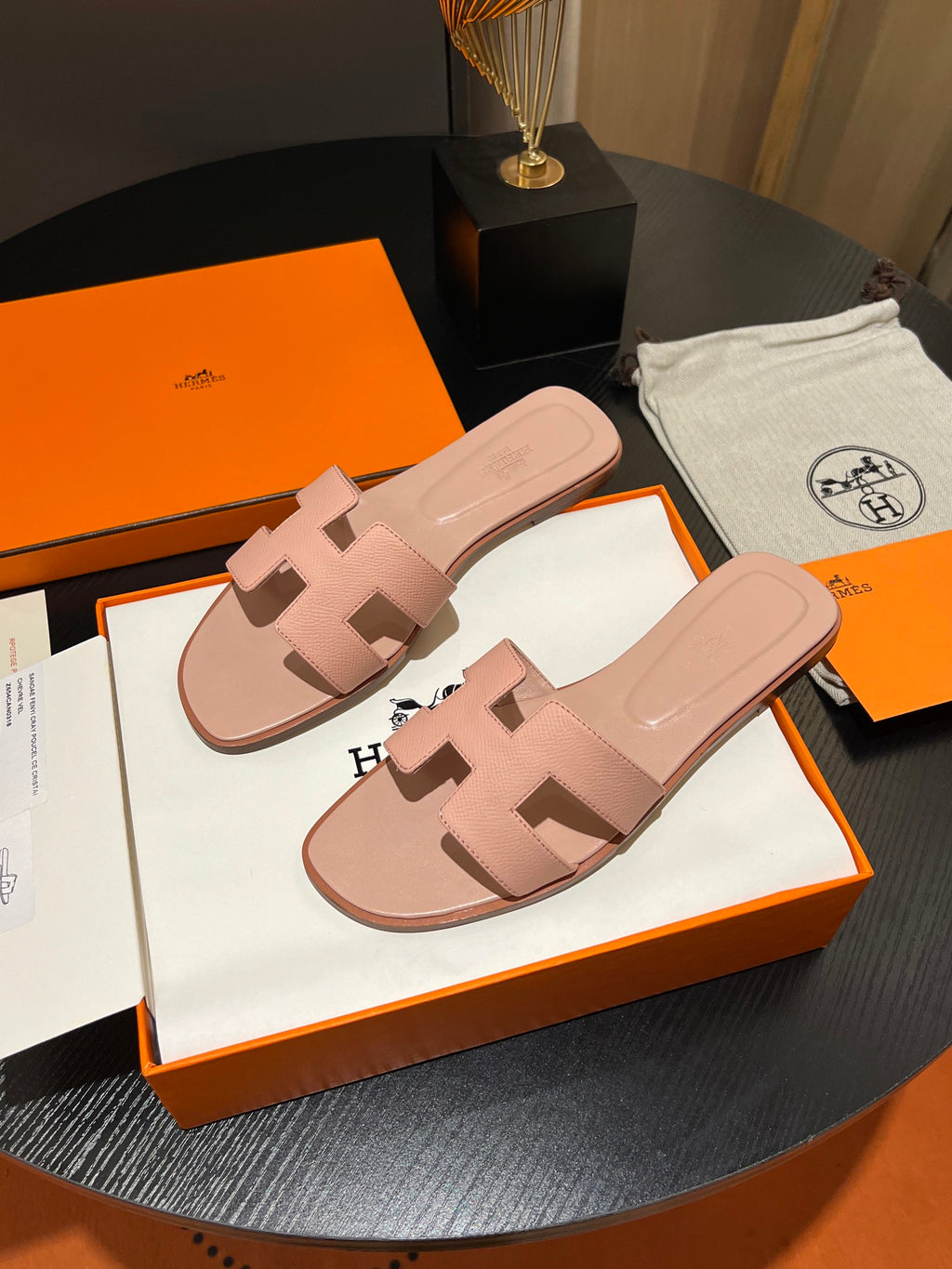 A pair of pink leather H-strap slide sandals by Hermès, displayed on their signature orange and white packaging boxes on a dark round table.