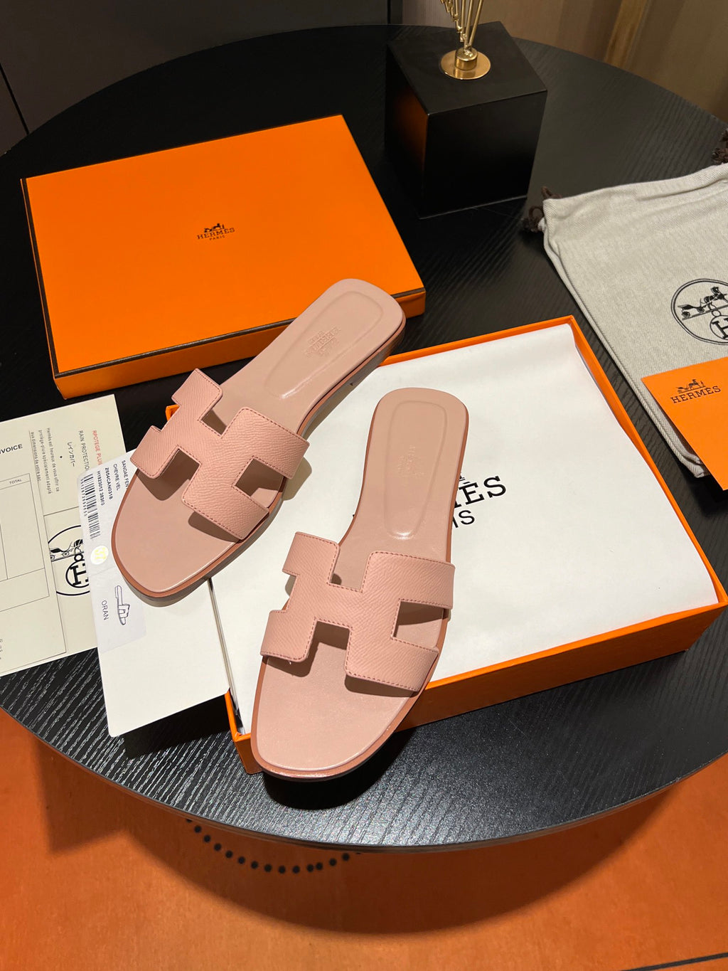 A pair of light pink leather slide sandals with an 'H' logo, displayed on top of an orange and white Hermes box on a round black table, with the orange Hermes box and dust bag in the background.