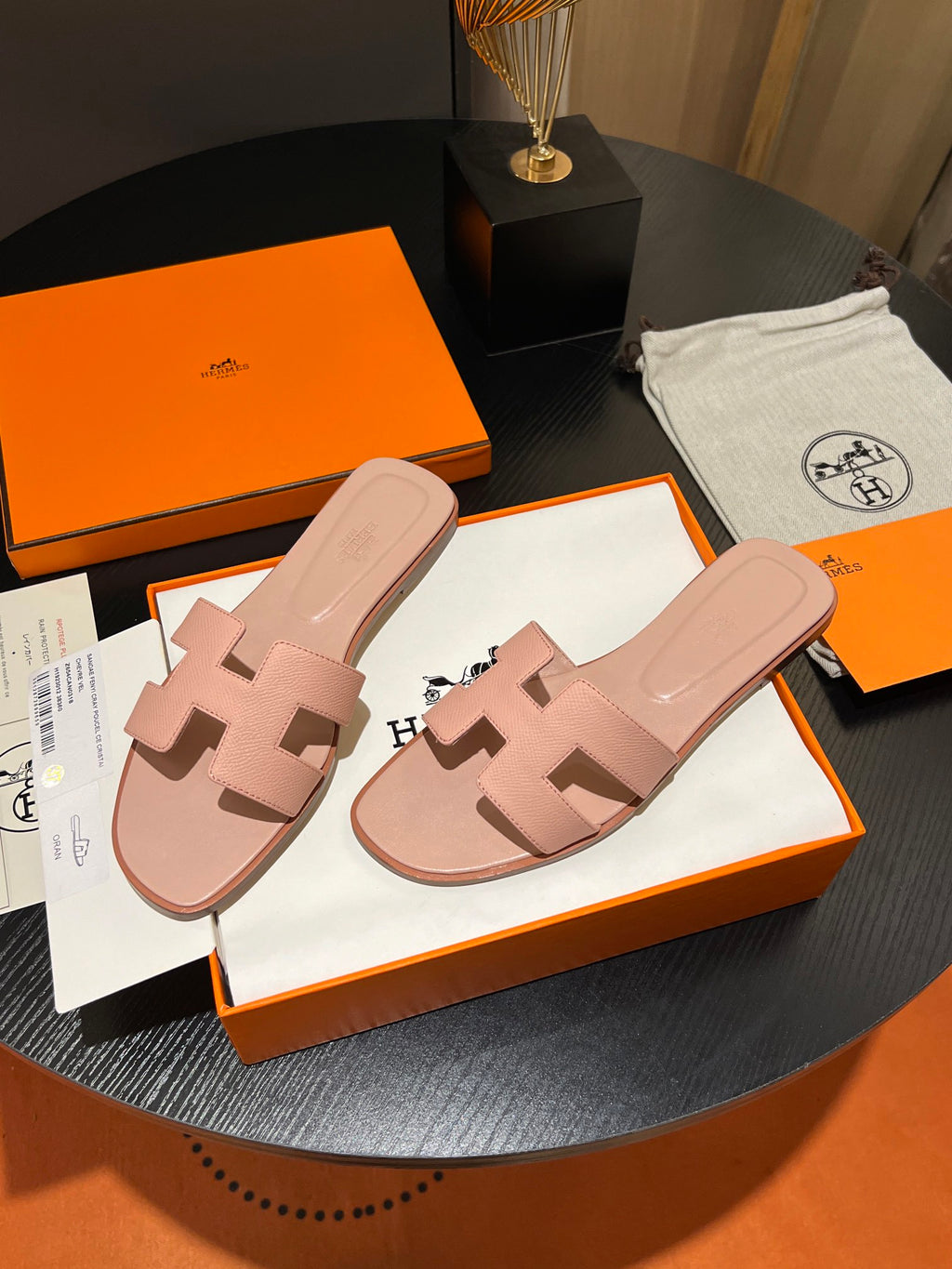A pair of pink leather H-strap slide sandals by Hermès, displayed on their signature orange and white packaging boxes on a dark round table.