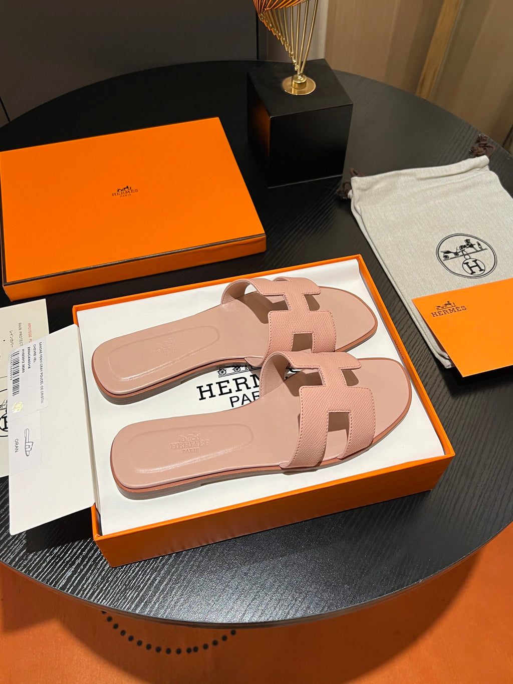 A pair of pink Hermès slide sandals in their original orange and white Hermès box, displayed on a dark round table alongside the orange box, dust bag, and papers.