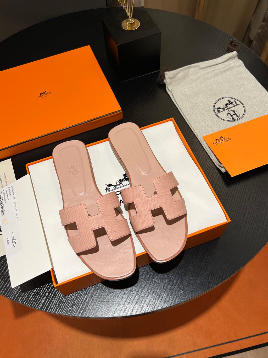 A pair of Hermès H-strap slide sandals in a dusty pink color, presented on their original orange and white Hermès boxes with a branded dust bag on a dark round table.