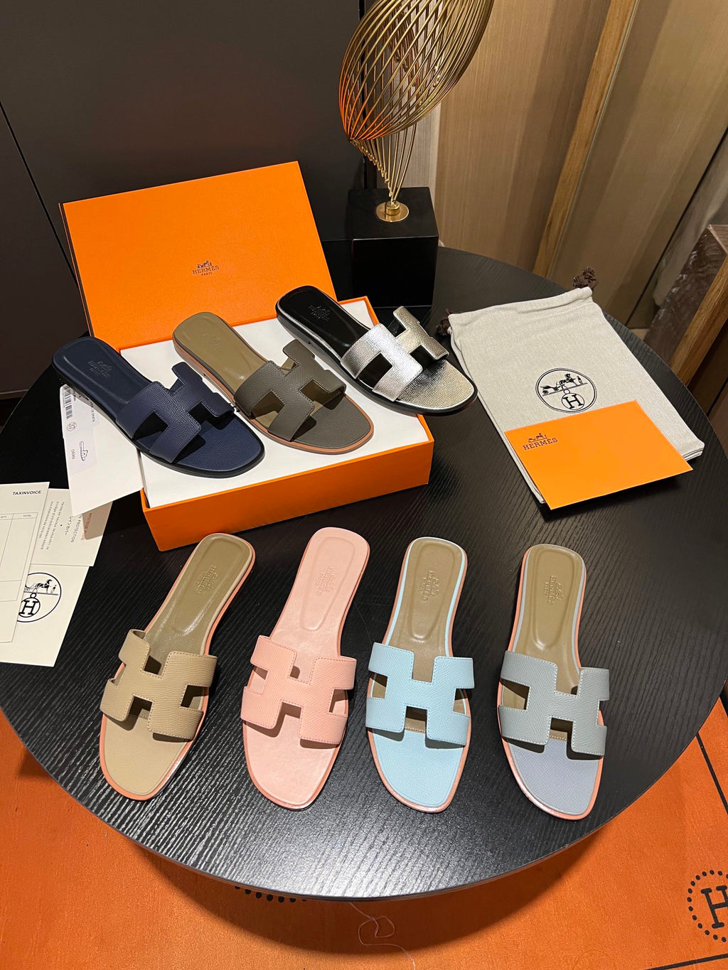 Four pairs of Hermès H-strap slide sandals in navy, taupe, white, beige, pink, light blue, and grey, displayed on a round black table alongside orange Hermès boxes and branded dust bags.