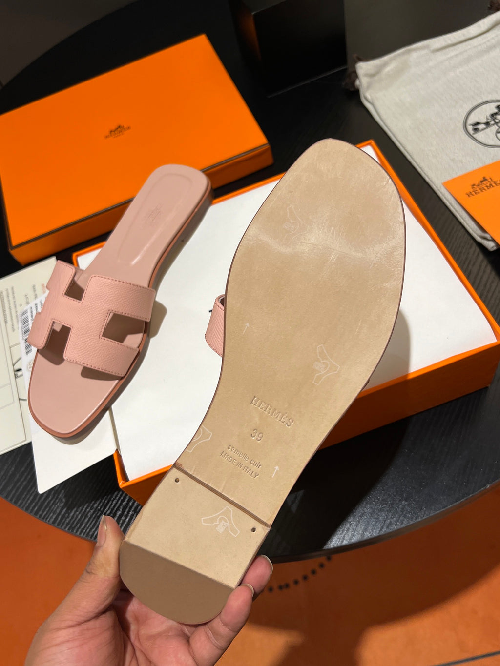 A hand holds the beige sole of a pink leather slide sandal, with another pink sandal and orange and white Hermès boxes in the background.
