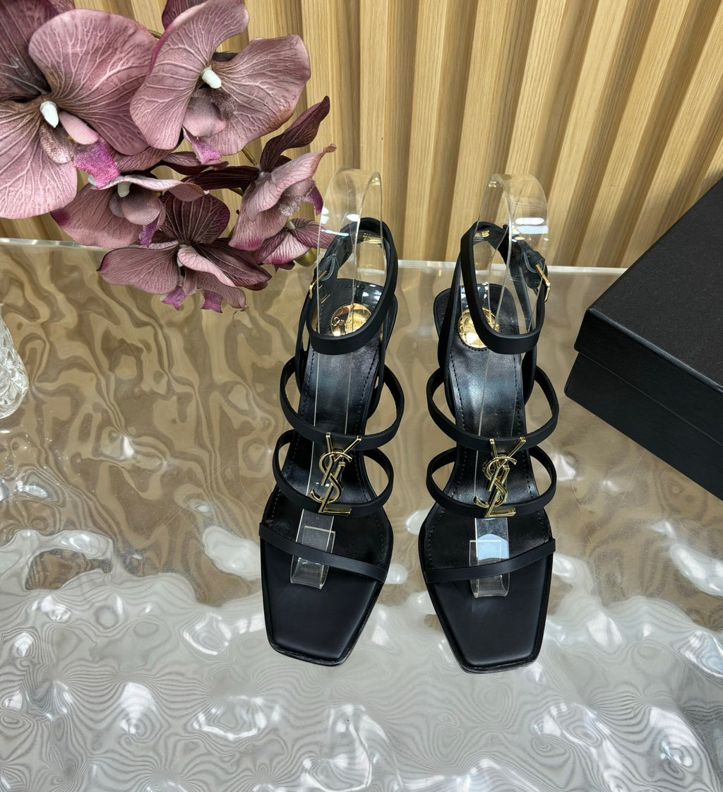 Black high-heeled sandals with clear straps on a reflective surface with flowers and a box in the background.