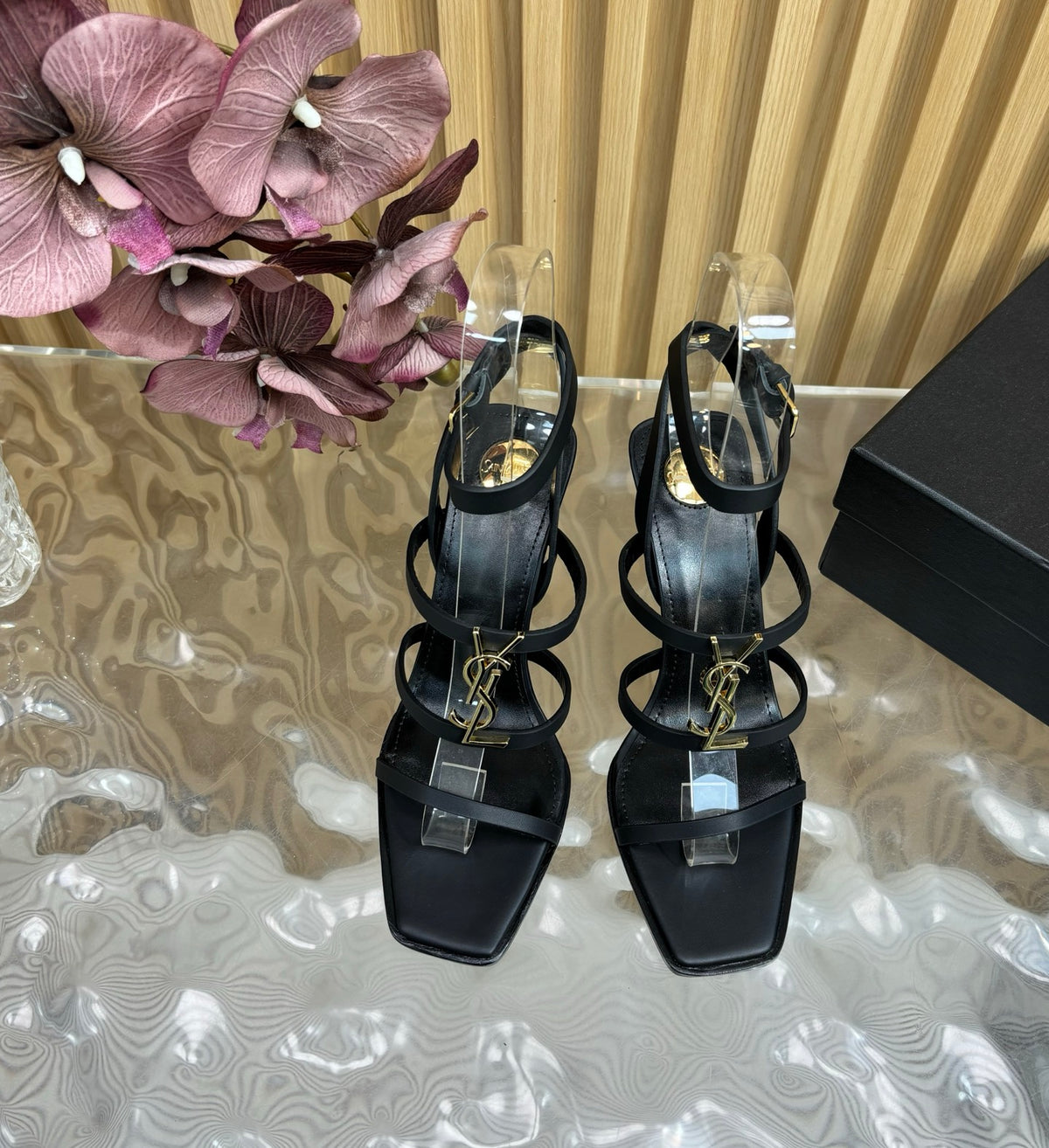 Black high-heeled sandals with clear straps on a reflective surface with flowers and a box in the background.