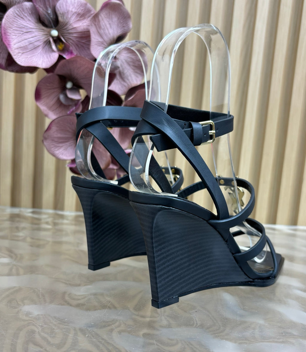 Black wedge sandals with clear straps on a textured surface with purple flowers in the background