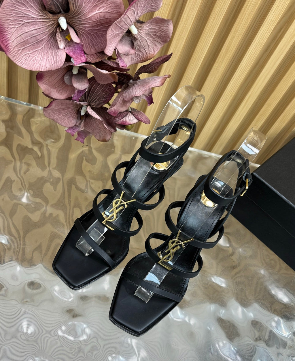 Black strappy sandals with gold accents on a marble surface with pink flowers in the background.