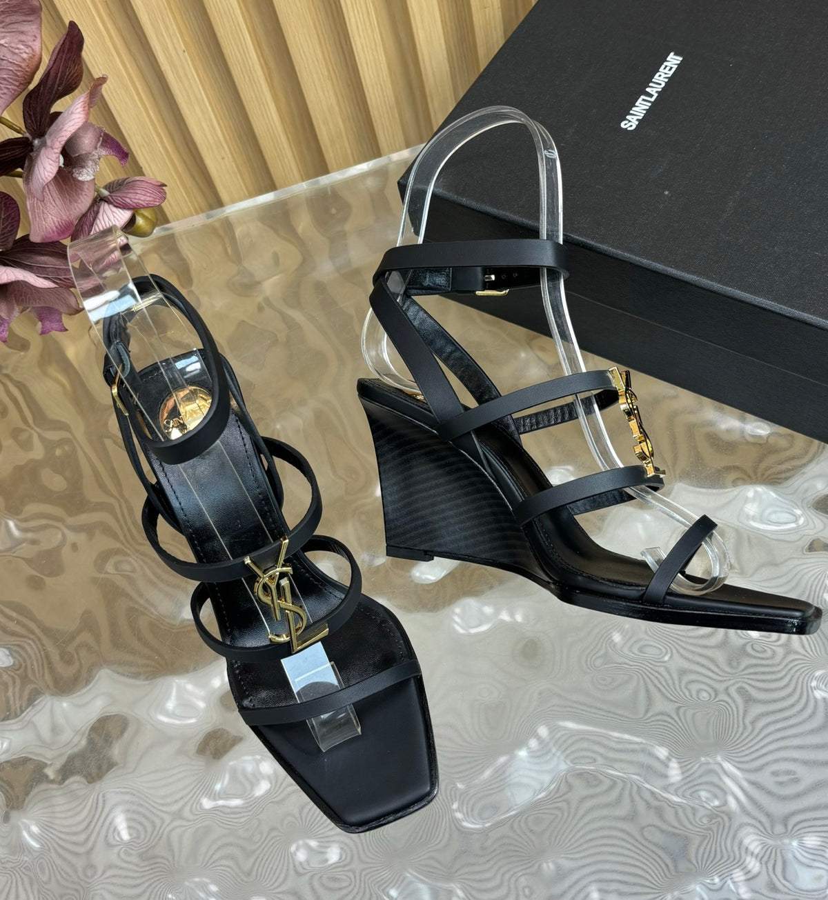 Black wedge sandals with gold accents on a reflective surface next to a black box.