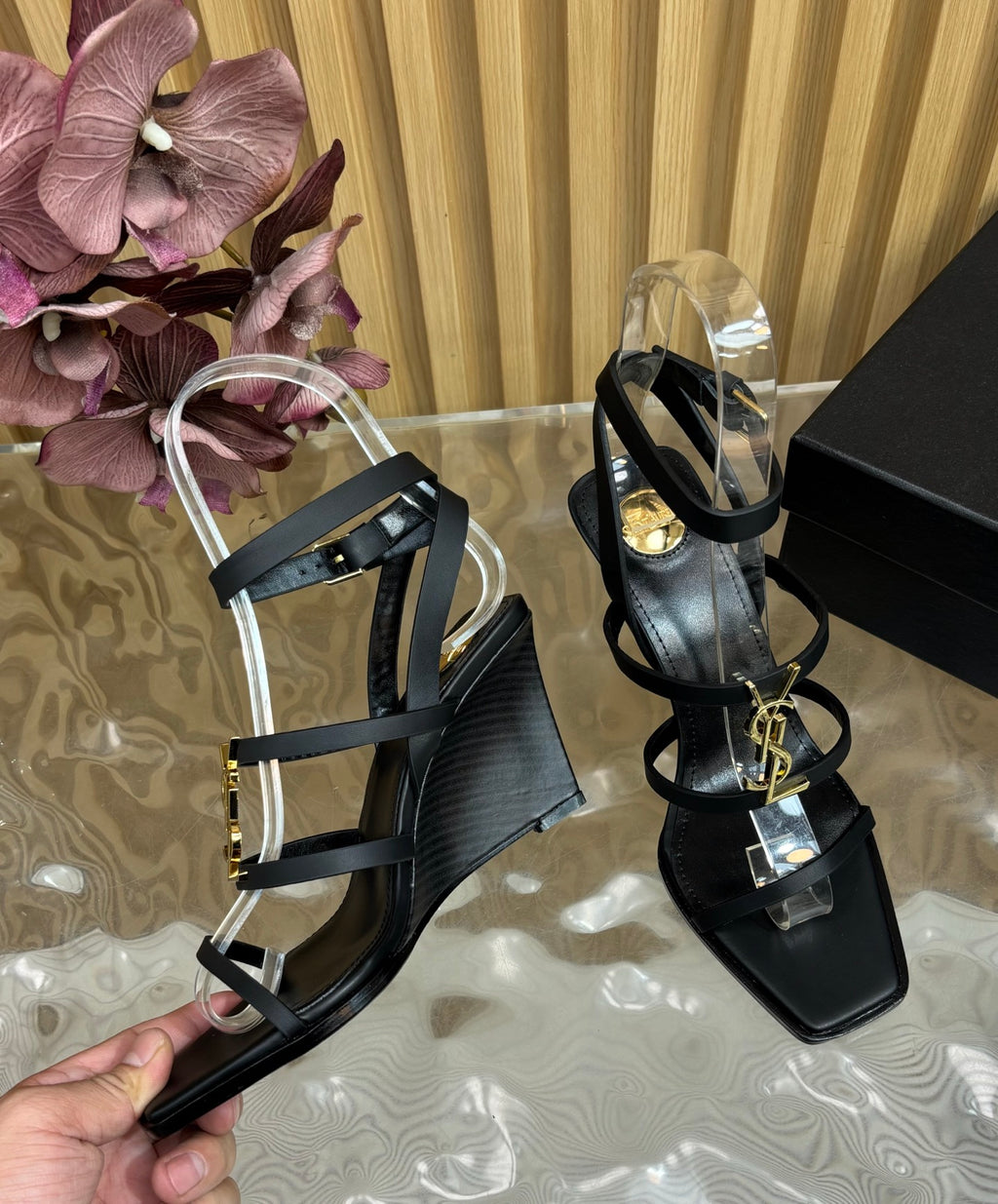 A pair of black strappy wedge sandals with clear acrylic heels and gold-tone hardware, shown on a reflective surface with a hand holding one sandal and a branded YSL box nearby.