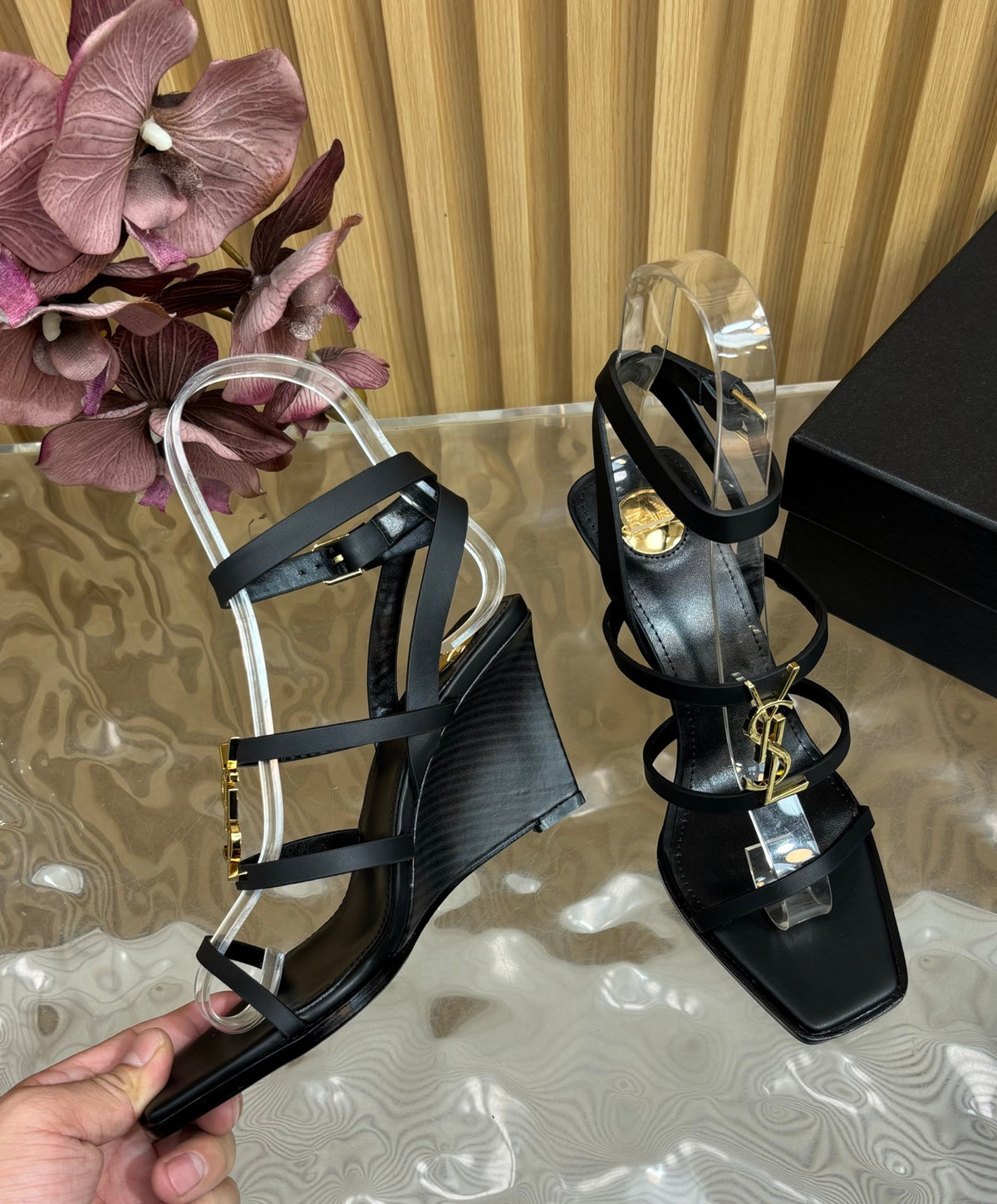 A pair of black strappy wedge sandals with clear acrylic heels and gold-tone hardware, shown on a reflective surface with a hand holding one sandal and a branded YSL box nearby.
